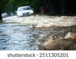 Close-up view of a flooded road during heavy rain, with a car driving through deep water in the background. The image captures the turbulent surface of the flood water