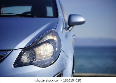 A close-up view of a front headhight lens. The blue of the sky and the sea in perfect harmony with the metallic color of the car.