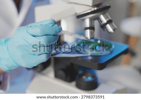 A close-up view of a gloved hand using a plastic dropper to add a liquid reagent into a petri dish placed on a microscope stage, illuminated by a cool blue light within the laboratory.