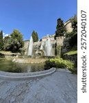 Close-up view of a grand fountain surrounded by lush greenery and historic architecture, with water cascading under a clear sky.