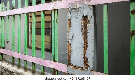 Close-up view of a green and pink iron house fence which is badly corroded due to rust spreading all over the iron surface.