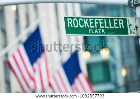 Close-up view of green street sign depicting it is Rockefeller Plaza in Midtown Manhattan, New-York. Blurred American flags in the background