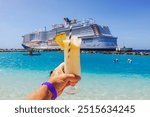 Close-up view of hand holding glass of alcoholic cocktail on Caribbean beach with cruise ship in background.