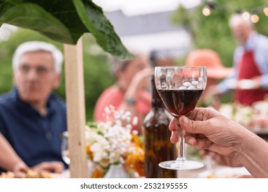 Close-up view of a hand holding a glass of red wine at an outdoor gathering of seniors. Blurred background shows elderly people, flowers, and a lakeside setting - Essence of a relaxed social event. - Powered by Shutterstock