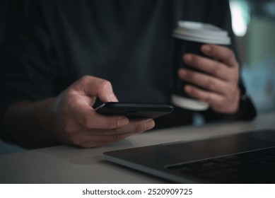 Close-up view of a hand holding a smartphone and a disposable coffee cup with a laptop on a desk, capturing a casual work environment. - Powered by Shutterstock