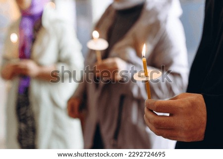 closeup view of hand with lit church candle in christian church, praying people with lit church candle in background, religion concept