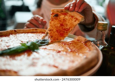 A close-up view of a hand serving a slice of hot, cheesy pizza with basil on top, alongside olive oil bottles at a cozy restaurant.