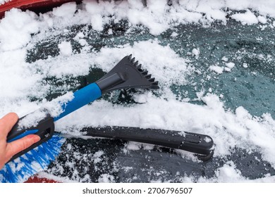 A close-up view of a hand using a winter scraper and brush to remove snow from a car windshield. The image highlights practical winter tasks, icy surfaces, and essential tools for cold-weather vehicle