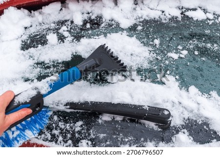 A close-up view of a hand using a winter scraper and brush to remove snow from a car windshield. The image highlights practical winter tasks, icy surfaces, and essential tools for cold-weather vehicle