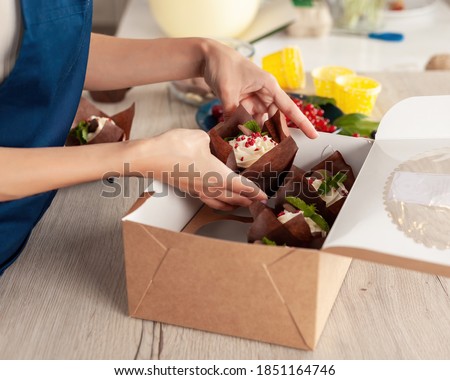 Closeup view of hands of female confectioner or baker in blue apron packing fresh decorated muffins with cream top to delivery craft box. Ready cupcakes with berries and mint in package.