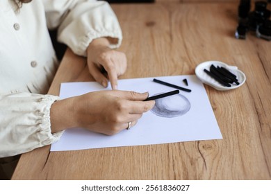Close-up view of hands skillfully drawing with charcoal on a blank paper, next to art supplies on a wooden surface