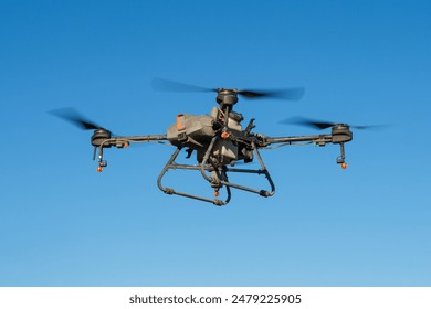 A closeup view of an herbicide sprayer agricultural drone flying in a clear blue sky. The drone has a large tank attached to its underside, and nozzles are visible on its arms. - Powered by Shutterstock