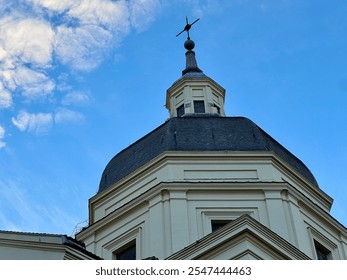 Close-up view of a historic church dome with a cross on top, set against a clear blue sky with some clouds. The architecture features classical details and symmetrical lines. - Powered by Shutterstock