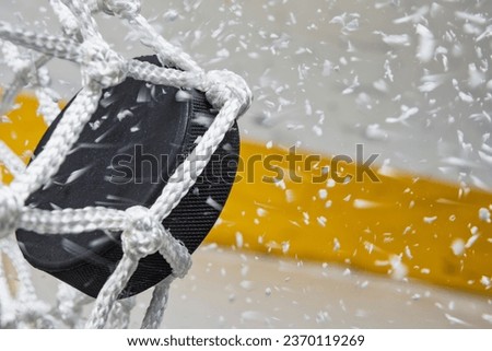 A close-up view of an Ice Hockey puck hitting the back of the goal net as shavings fly by, viewed from the side. Scoring a goal in ice hockey.