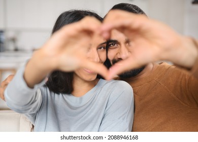 Close-up of view of the Indian couple of spouses looking at the camera and showing heart shape from the hands while sitting at the sofa at their new flat. Stock photo - Shutterstock ID 2088946168