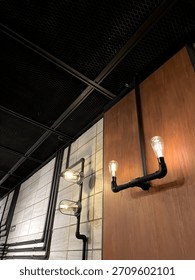 A close-up view of an interior space featuring industrial-style sconce lights. The wall is a combination of textured tiles and warm wood paneling, with exposed black pipe conduit. 