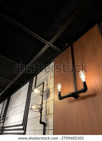 A close-up view of an interior space featuring industrial-style sconce lights. The wall is a combination of textured tiles and warm wood paneling, with exposed black pipe conduit. 