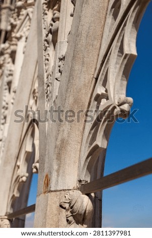 A close-up view of intricate Gothic stone tracery and carvings on the facade of the Duomo di Milano, set against a vibrant clear blue sky.