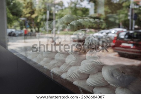 A close-up view of a large display of white, swirled meringue cookies in a bakery or cafe window. The glass window has strong, clear reflections of an outdoor street scene.