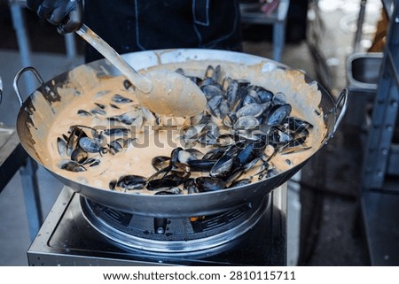Close-up view of a large shallow pan filled with black-shelled mussels being cooked in a rich creamy saffron-colored sauce. A gloved hand holds a long-handled ladle, stirring the