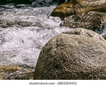 Close-up view of a large, solid grey river boulder with white frothy stream water cascading behind it.
