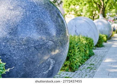 Close-up view of large spherical stone sculptures arranged in a row along a paved walkway in an outdoor urban park setting. The foreground features a massive dark gray granite sphere with visible natural stone texture, surface cracks, and mineral patterns. Additional similar stone balls recede into the background with shallow depth of field, creating a perspective line. Lush green hedges and bushes grow between the sculptures and the cobblestone pathway. The scene is illuminated by bright daylight with dappled sunlight filtering through trees in the blurred background. The composition emphasizes the contrast between the smooth geometric round shapes of the stone sculptures and the organic vegetation surrounding them.