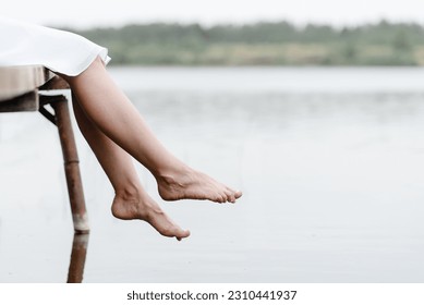 closeup view of legs, young beautiful caucasian woman in short white dress sits at wooden pier on river bank in summer, feet dangled in water, green trees in background, melancholy concept, copy space - Powered by Shutterstock