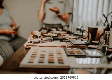 A close-up view of a makeup artist’s table featuring eyeshadow palettes, brushes, and beauty products with blurred figures in the background, highlighting a preparation scene for an event.