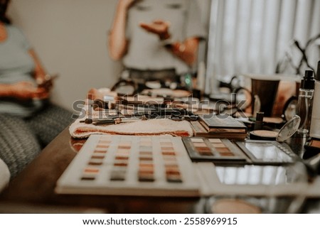 A close-up view of a makeup artist’s table featuring eyeshadow palettes, brushes, and beauty products with blurred figures in the background, highlighting a preparation scene for an event.
