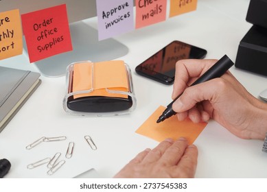 closeup view of a man about to write with a black marker on a blank orange sticky note on a clean office workspace, surrounded by stationery, and colorful task notes on a monitor