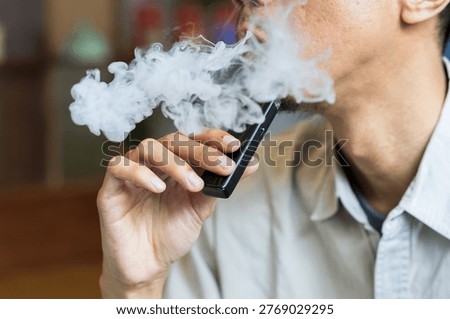 Close-up view of a man actively vaping with a modern, black pod-style electronic cigarette. A man holds a vape device while exhaling thick white vapor curls around his hand and mouth.