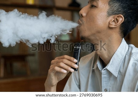 Close-up view of a man actively vaping with a modern, black pod-style electronic cigarette. A man holds a vape device while exhaling thick white vapor curls around his hand and mouth.