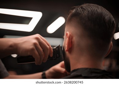 A close-up view of a man getting his beard trimmed at a barbershop. The barber’s hand holds electric clippers near the client’s face, showcasing grooming precision under modern lighting.
