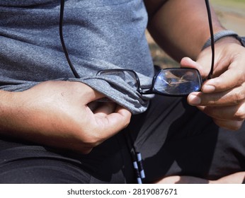 A close-up view of a man's hands using the hem or fabric edge of his grey t-shirt to wipe and clean his eyeglasses outdoors