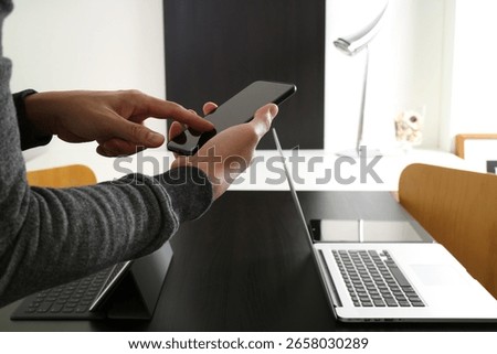  A close-up view of a man's hands using a smartphone in a modern office setting with a laptop and tablet on a black wooden desk, representing technology and digital communication.