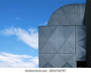 A close-up view of the metallic, angular exterior of a building against a bright blue sky with scattered clouds. The building's facade features geometric patterns formed by riveted metal panels.