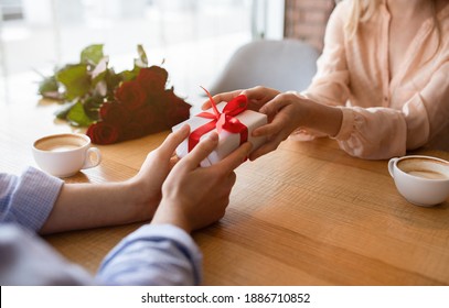 Closeup view of millennial guy giving his girlfriend Valentine's or birthday gift at table in cozy cafe. Unrecognizable young couple celebrating special occasion together, exchanging presents