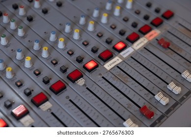 A close-up view of a mixing board with multiple knobs, sliders, and lights in a recording studio. The equipment is in use during a music session in the afternoon.