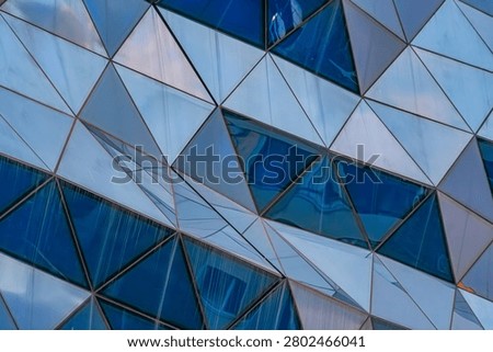 Close-up view of modern architectural facade, featuring geometric triangular glass panels, interplay of light and shadow, reflections of urban environment, structured composition, cool color tones