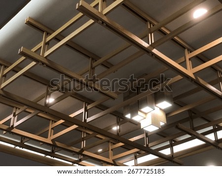A close-up view of a modern ceiling with a complex grid of light brown beams. The geometric pattern is accented by several recessed lights and a central hanging lamp, giving the space a clean
