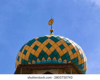 A close-up view of a mosque's dome, featuring a distinctive pattern of yellow and teal tiles, topped with a golden finial, set against a vibrant blue sky.