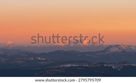 Close-up view of mountain range in Carpathians illuminated by warm light of sunset. Snow-dusted peak, orange sky, foothills covered in forest and snow, atmospheric mood, serene evening landscape.