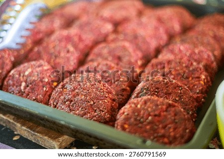 A close-up view of multiple raw meat patties arranged in neat rows inside a rectangular metal or plastic tray. The patties are round, reddish in color, and generously seasoned with visible spices and herbs. A white plastic serving utensil with serrated edges is partially visible in the upper left corner. The meat appears to be ground and formed into uniform circular shapes, likely prepared for grilling or frying. The texture of the meat is coarse with visible bits of fat and seasoning throughout. The setting suggests a butcher shop, market stall, or food preparation area. The focus is selective, with the foreground patties in sharp detail while the background gradually blurs.