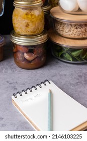 Closeup View Of A Notepad With A Pencil Over It In Front Of Several Piles Of Prepared Meal Glass Containers.