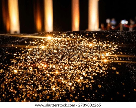 Close-up view of numerous small, sparkling golden glitter particles scattered across a dark, textured surface. Softly lit, out-of-focus columns are visible in the background, creating a bokeh effect.