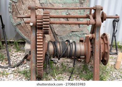 A close-up view of an old, rusty mechanical winch featuring large metal gears, a coiled rope, and weathered components. The corroded surface and aged texture highlight industrial deca