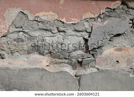 A close-up view of an old wall displaying peeling paint and crumbling plaster, revealing various textures and colors. The urban environment shows the wear and tear of time in broad daylight