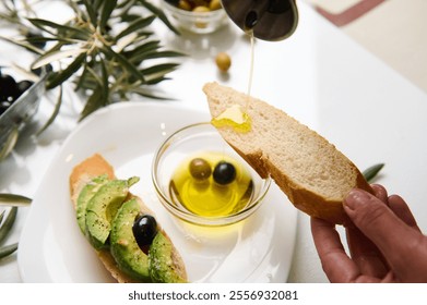 A close-up view of olive oil being poured on crusty bread, with avocado toast and olives on a table. - Powered by Shutterstock