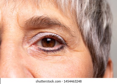 A Closeup View On The Eye Of An Old-aged Woman Waring Blue Eye Shadow And Eyeliner. Lady With Brown Eyes And Grey Hair With Wrinkled Face.