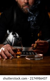close-up view on hand of a male bartender holding transparent glass with cognac and smoking cuban cigar over ashtray on table - Shutterstock ID 2174986437